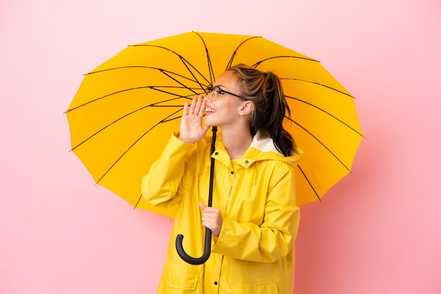 Teenager Russian girl with rainproof coat and umbrella isolated on pink background shouting with mouth wide open to the side