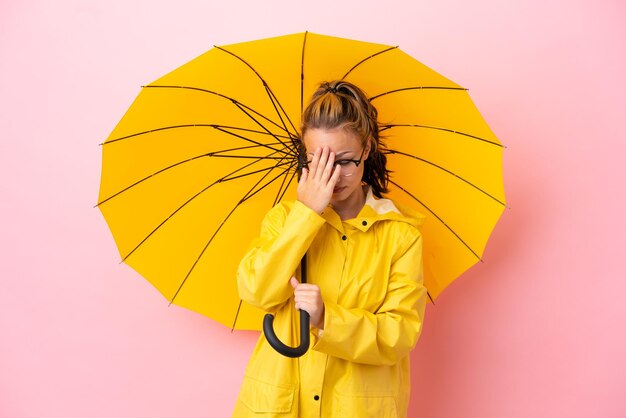 疲れたと病気の表情でピンクの背景に分離された防雨コートと傘を持つ10代のロシアの女の子