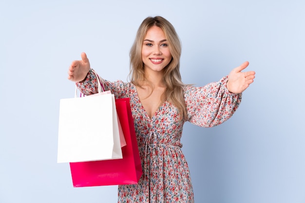 Teenager Russian girl with shopping bag isolated on blue wall presenting and inviting to come with hand