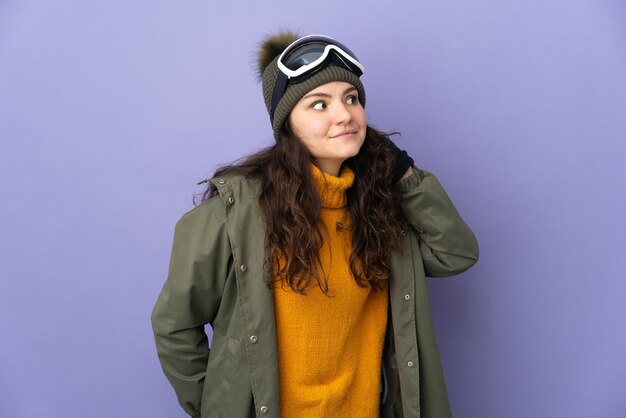 Teenager Russian girl with snowboarding glasses isolated on purple background listening to something by putting hand on the ear