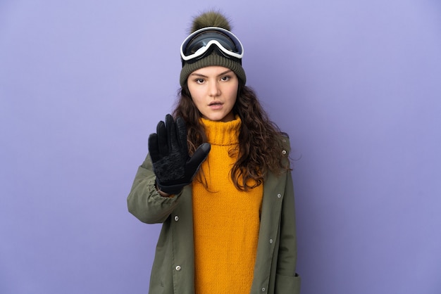 Teenager Russian girl with snowboarding glasses isolated on purple background making stop gesture