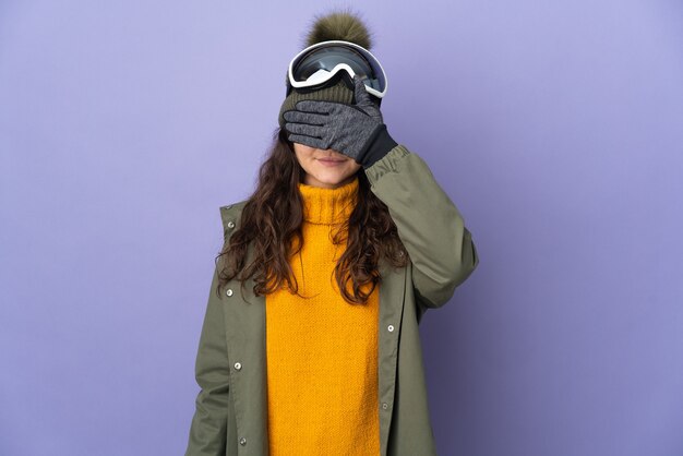 Русская девушка подростка в очках сноуборда, изолированных на фиолетовой поверхности, закрывающей глаза руками. Не хочу что-то видеть