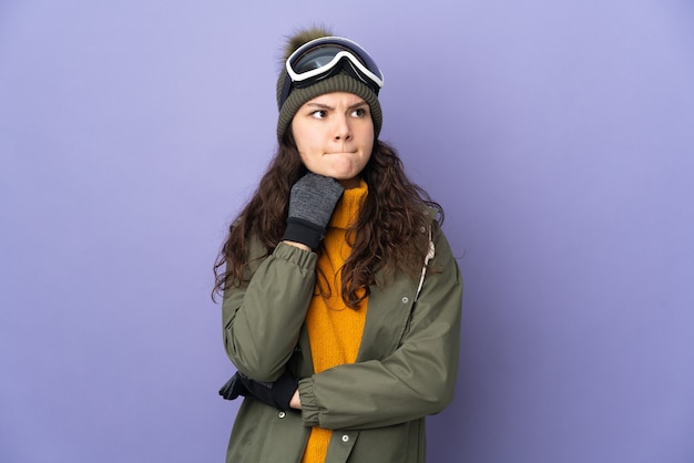 Teenager Russian girl with snowboarding glasses isolated on purple wall having doubts and thinking