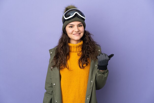 Teenager Russian girl with snowboarding glasses isolated on purple wall pointing to the side to present a product