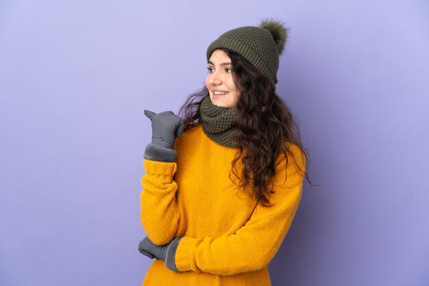 Фото Русская девушка-подросток в зимней шапке изолирована на фиолетовой стене, указывая в сторону, чтобы представить продукт