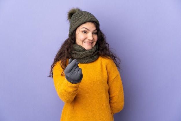 Ragazza russa dell'adolescente con il cappello di inverno isolato su priorità bassa viola che fa gesto dei soldi