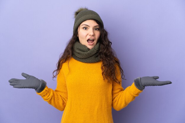 ショックを受けた表情で紫色の背景に隔離された冬の帽子を持つ 10 代のロシアの女の子