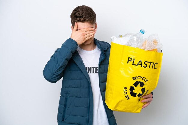 Teenager Russian holding a bag full of plastic bottles to recycle on white background covering eyes by hands Do not want to see something