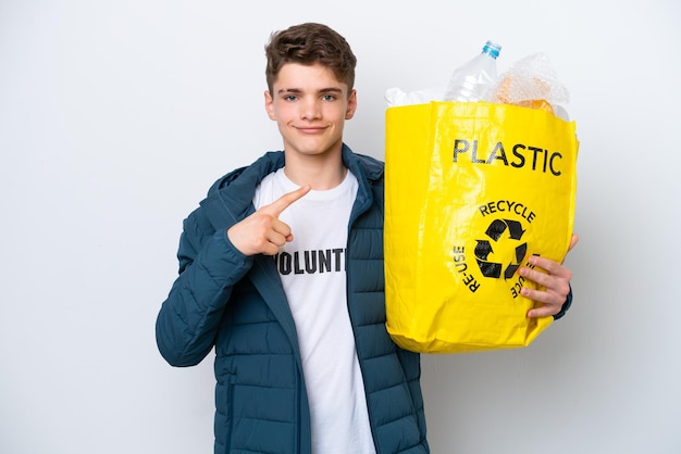 Teenager Russian holding a bag full of plastic bottles to recycle on white background pointing to the side to present a product