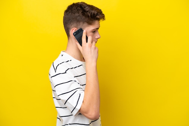 Teenager Russian man isolated on yellow background keeping a conversation with the mobile phone with someone