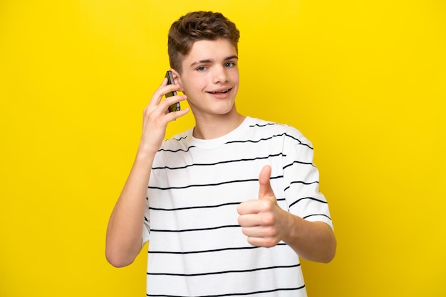 Teenager Russian man isolated on yellow background keeping a conversation with the mobile while doing thumbs up