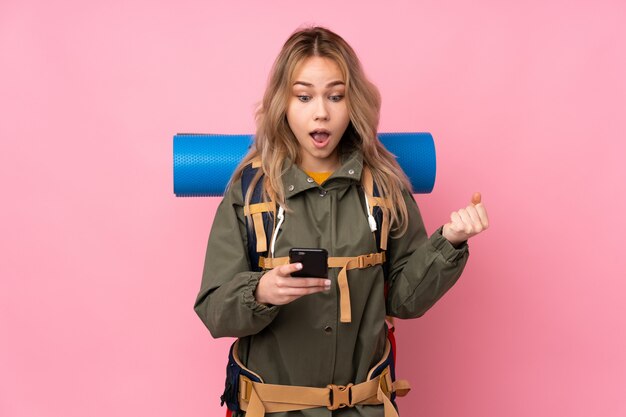 Teenager Russian mountaineer girl with a big backpack on pink wall surprised and sending a message