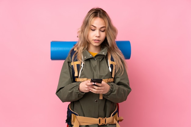 Teenager Russian mountaineer woman with a big backpack isolated on pink wall sending a message with the mobile