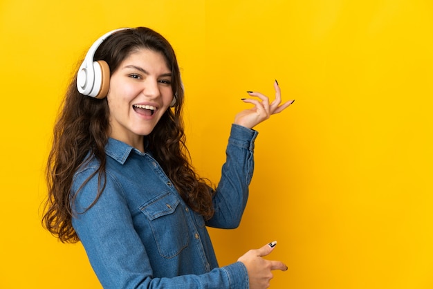 Teenager Russian woman isolated on yellow wall listening to music and doing guitar gesture