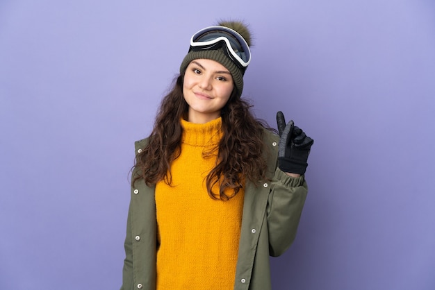 Teenager russian woman with snowboarding glasses isolated on
purple wall showing and lifting a finger in sign of the best