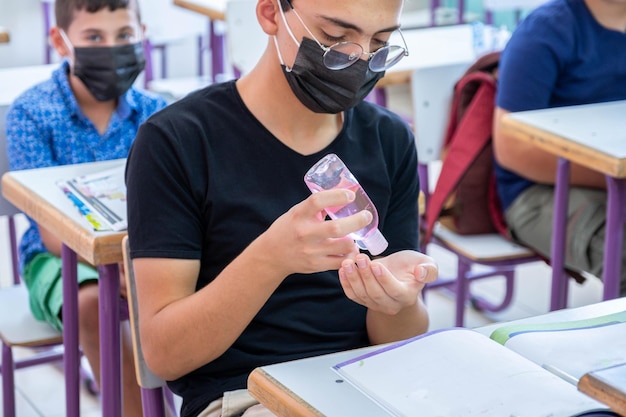 Teenager in school sterilizing his hands while wearing face mask to prevent infection