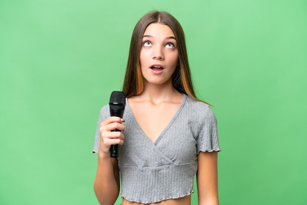 Teenager singer girl picking up a microphone over isolated background looking up and with surprised expression