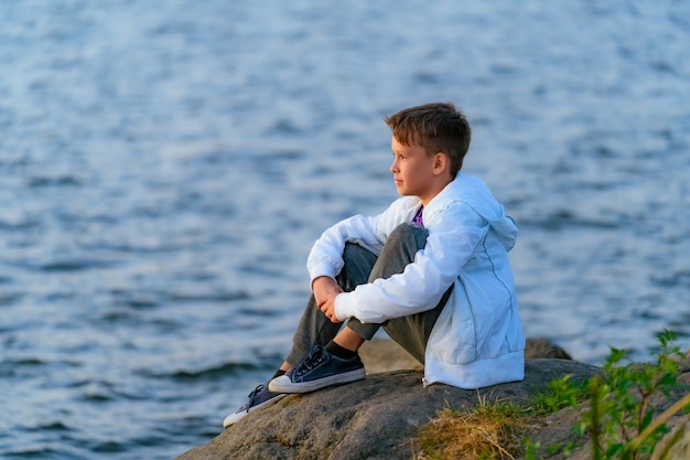 The teenager sits a large stone boulder on the bank of the river and looks at the river below. Summer vocation. Bright green vegetation, blue sky.