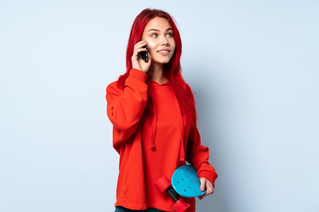 Teenager skater woman isolated on white wall keeping a conversation with the mobile phone