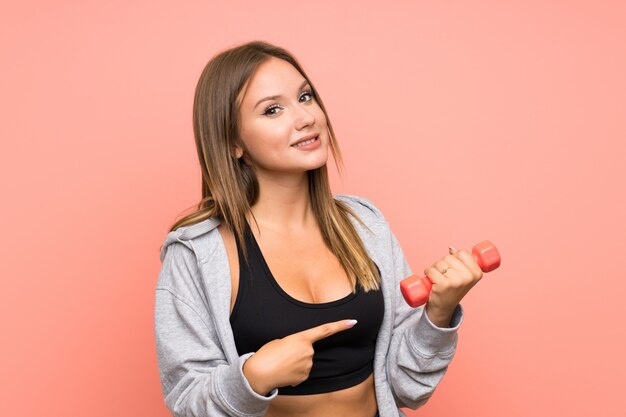 Ragazza di sport dell'adolescente che fa sollevamento pesi sopra la parete rosa isolata e che lo indica