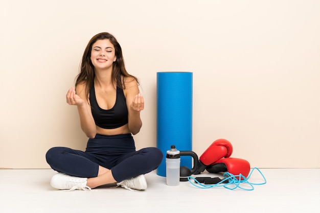 Ragazza di sport dell'adolescente che si siede sul pavimento che fa gesto dei soldi