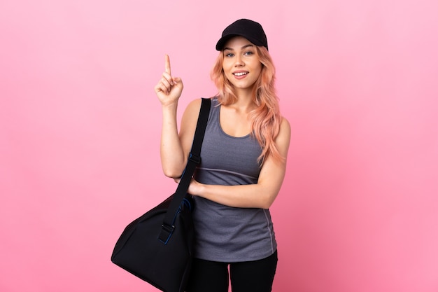 Teenager sport woman with sport bag over isolated wall happy and pointing up