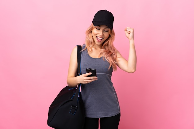 Подростковая спортивная женщина со спортивной сумкой над изолированной стеной с телефоном в победной позиции