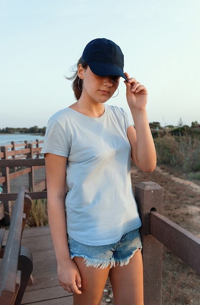 사진 해질녘 바다 옆에 서 있는 10대. 티셔츠와 짙은 파란색 야구 모자를 쓰고 바이저를 만지는 십대 소녀. 그늘에서 얼굴. 모자와 티셔츠 모형