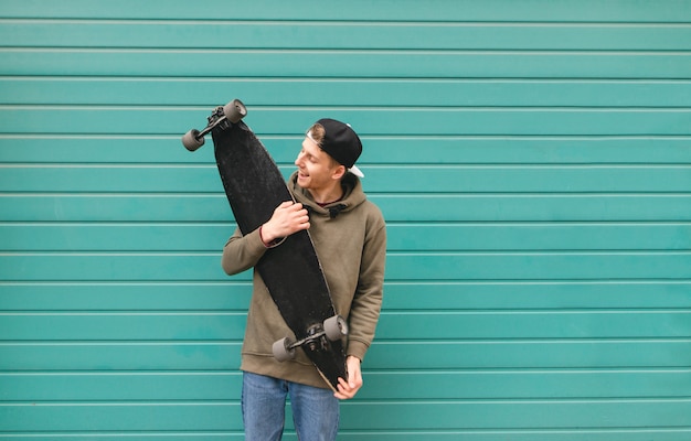 Teenager in street clothes standing with a longboard in his hands on a turquoise background