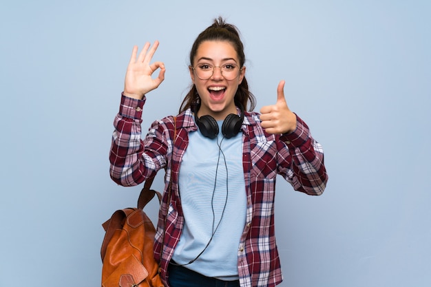 Teenager student girl over isolated blue wall showing ok sign and thumb up gesture