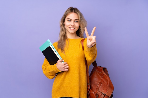 Teenager student girl on purple wall smiling and showing victory sign