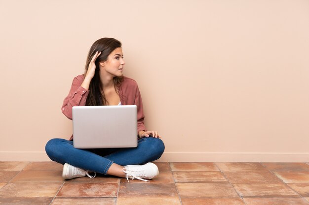 Teenager student girl sitting on the floor with a laptop listening to something by putting hand on the ear