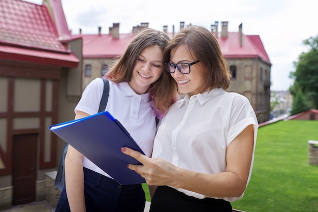 Teenager student girl talking with teacher, woman with clipboard reading and writing in paper document from outside the school. Back to school, back to college, education concept