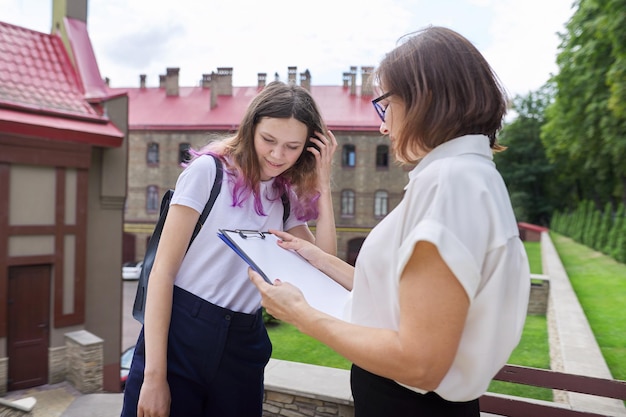 Teenager student girl talking with teacher, woman with clipboard reading and writing in paper document from outside the school. Back to school, back to college, education concept