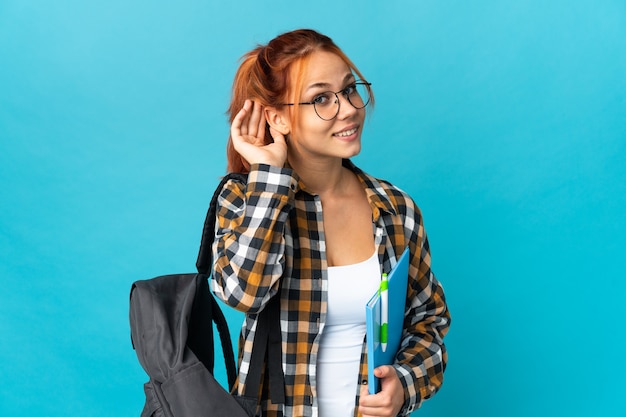 Teenager student Russian girl isolated on blue wall listening to something by putting hand on the ear