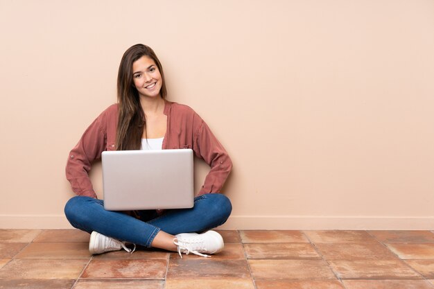 Teenager student woman sitting on the floor with a laptop posing with arms at hip and smiling