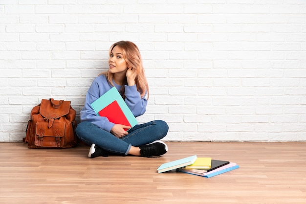 ピンクの髪の10代の学生女性が屋内で床に座って耳に手を置いて何かを聞いている