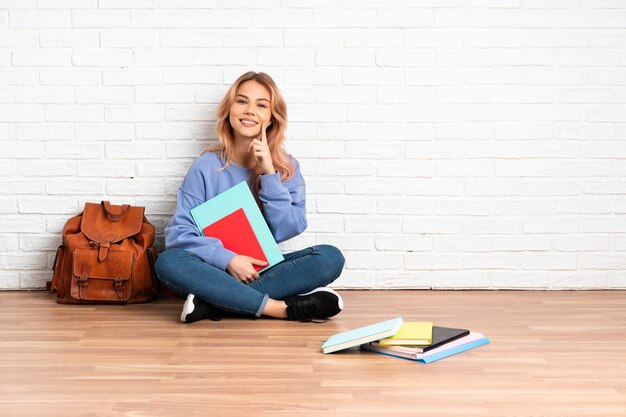 Teenager student woman with pink hair sitting on the floor at indoors smiling with a happy and pleasant expression