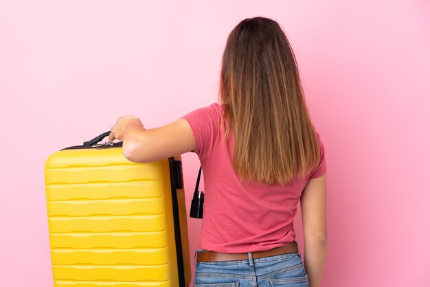 Teenager traveler woman holding a suitcase over isolated pink wall in back position