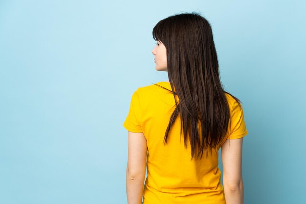 Teenager Ukrainian girl isolated on blue background in back position and looking side