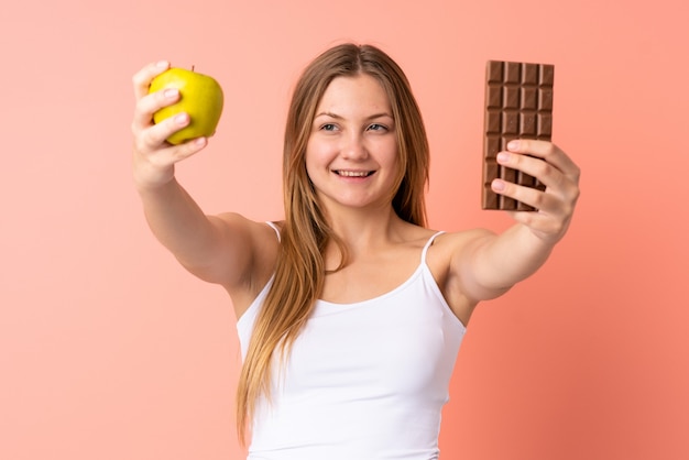 Teenager Ukrainian girl isolated on pink taking a chocolate tablet in one hand and an apple in the other