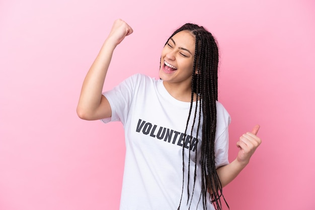 Teenager volunteer girl with braids isolated on pink background celebrating a victory