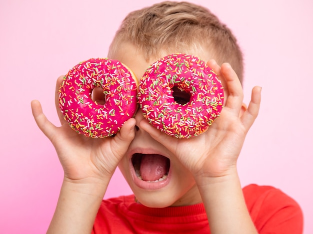 The teenager was having fun with donuts looking through them. a boy is having fun with donuts on a pink background. fun time with food