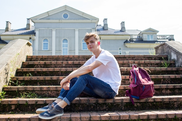 teenager in a white T-shirt with a school backpack sits on the steps in the park