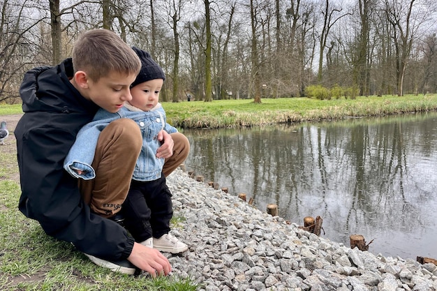 Photo a teenager with a baby brother are watching birds by the river in early spring