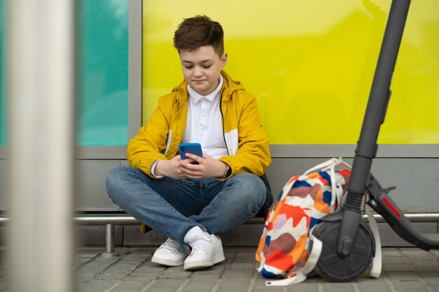 Подросток с мобильным телефоном сидит рядом со своим электросамокатом