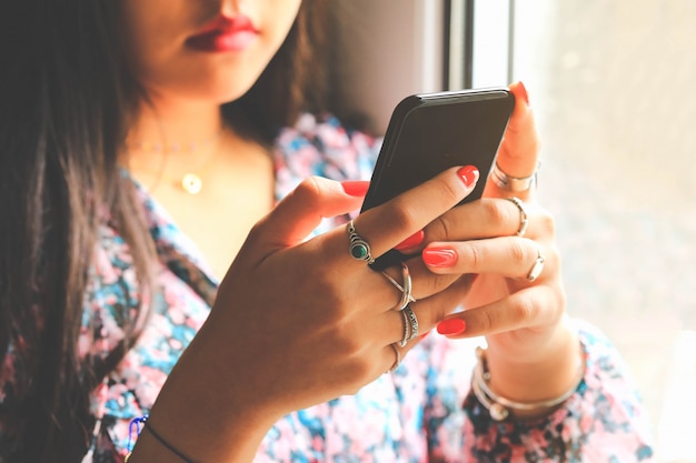 Adolescente con le unghie rosse del manicure facendo uso dello smartphone moderno vicino alla finestra