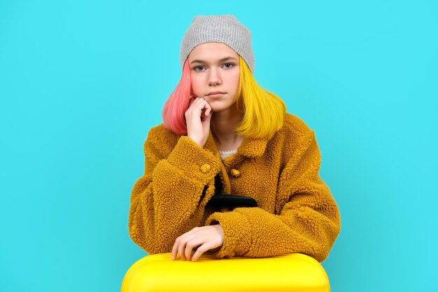 Teenager woman in hat fur coat with suitcase on blue turquoise background. Vacation, study, tourism winter holidays concept