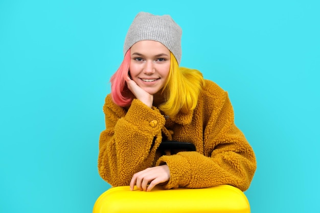 Teenager woman in hat fur coat with suitcase on blue turquoise background. Vacation, study, tourism winter holidays concept