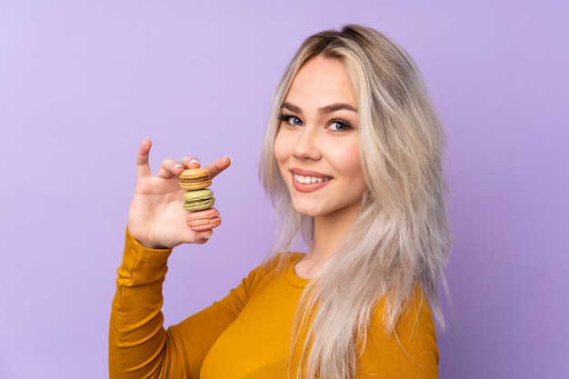 Teenager woman over isolated purple wall holding colorful French macarons and smiling a lot